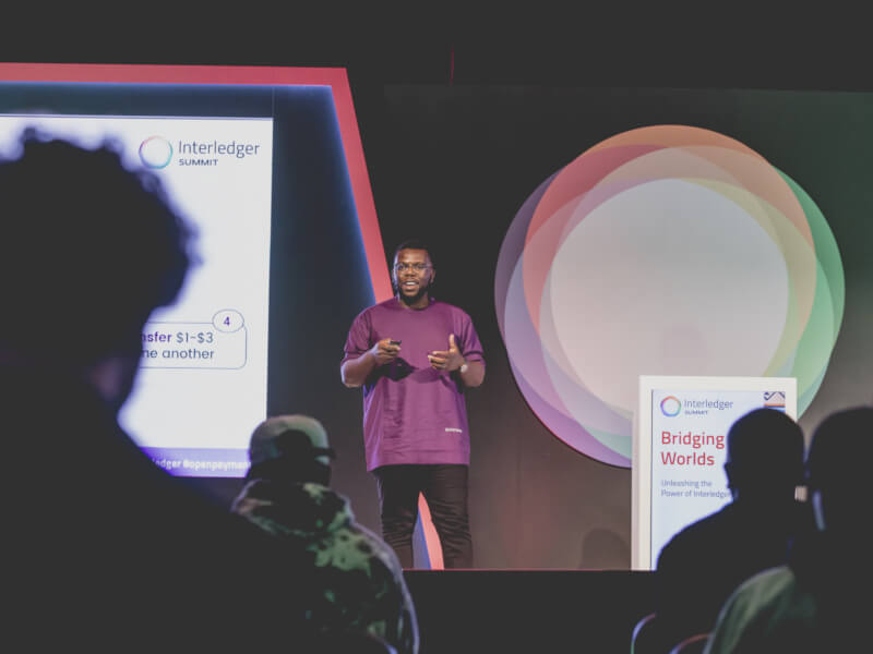 Uchi Uchebeke stands on stage giving a presentation at an Interledger Summit, with audience silhouettes visible in the foreground between him and the camera.